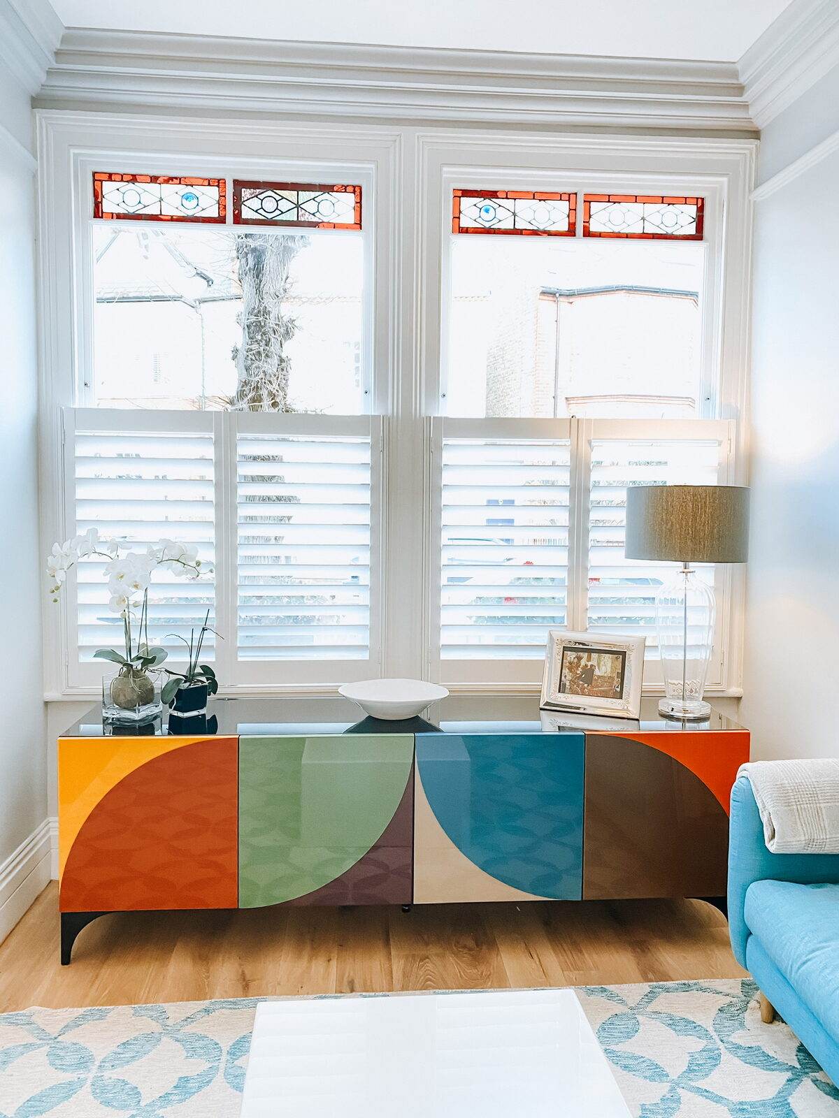 White shutters across a bay window above a colourful sideboard