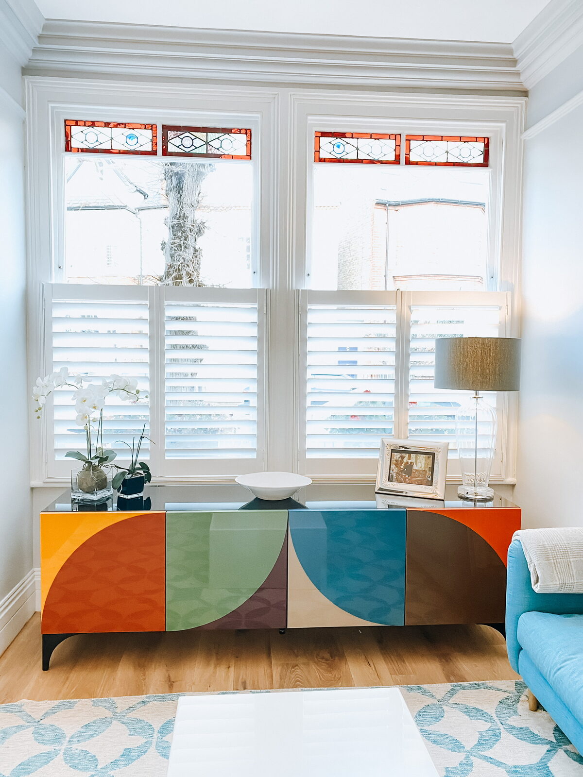 White shutters across a bay window above a colourful sideboard